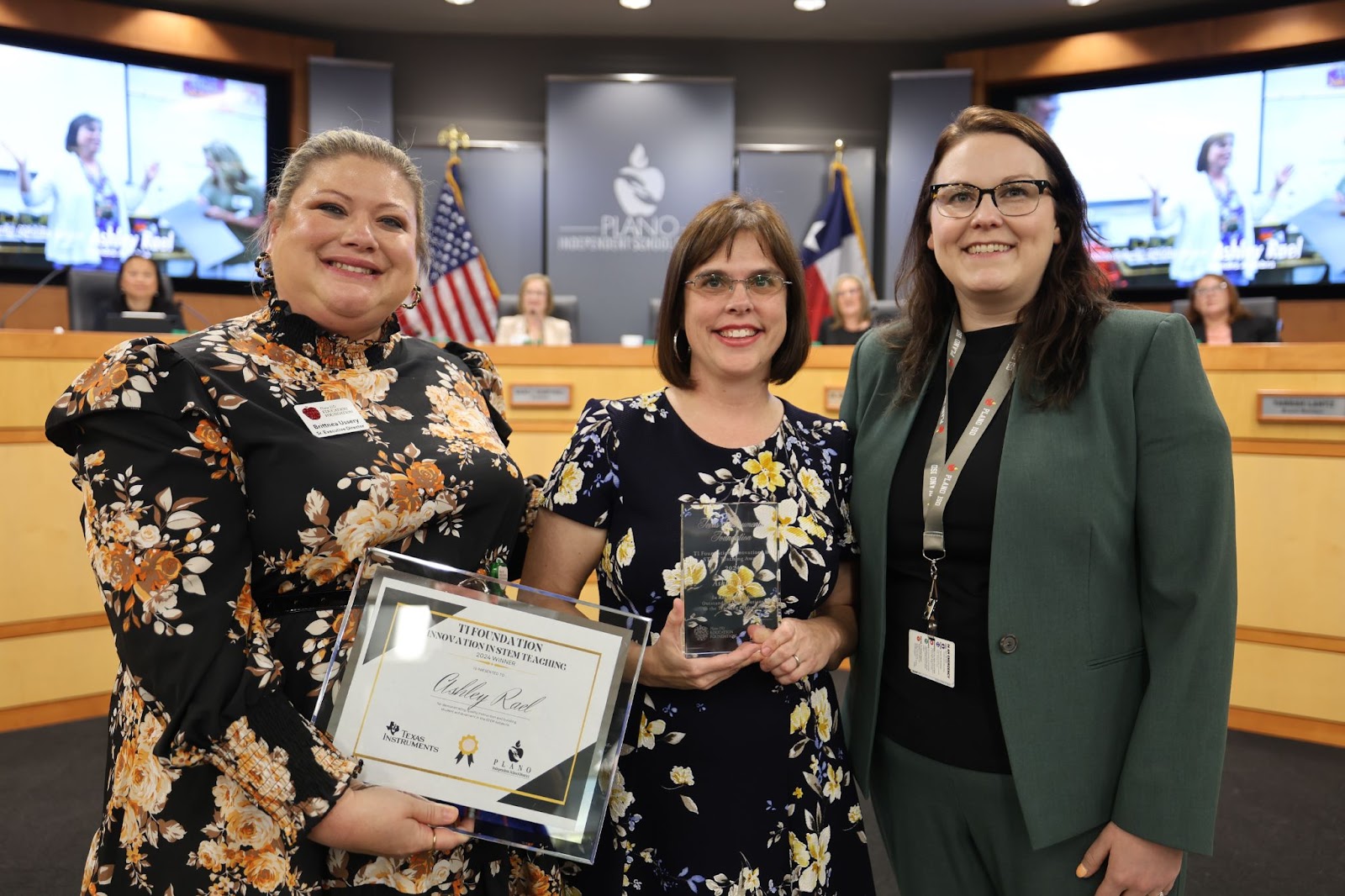 three individuals standing in front of the Plano ISD boardroom, the middle woman is holding a certificate and plaque